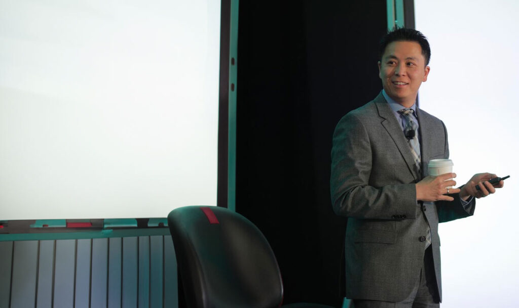 Dr. Samson Liu speaking at a professional event, holding a coffee cup while presenting leadership and dental practice growth strategies.